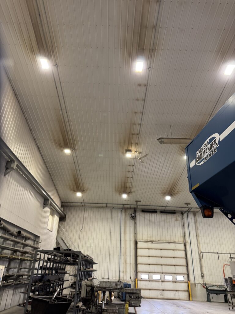 Hydronic-heated ag shop ceiling showing dark grime streaks running between LED fixtures along the steel panel ribs, with a stained overhead door visible below.